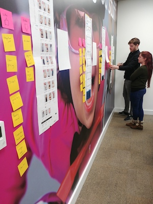 Two people chatting while looking at a web page which has been printed and stuck on the wall.